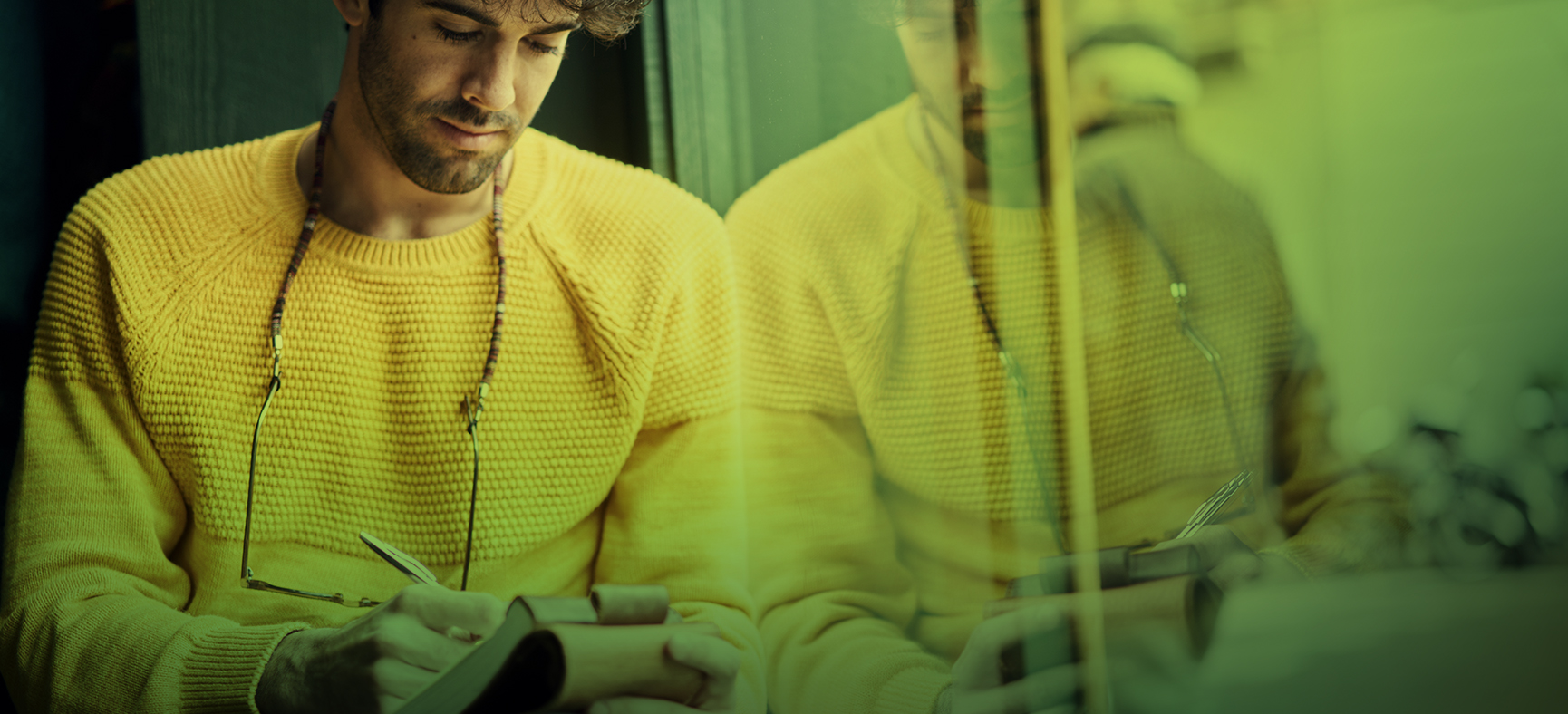 Man leans against a window with his reflection while he writes in a notepad